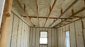 A room's interior with spray foam insulation on walls and ceiling, exposed wooden beams, and two windows covered with plastic sheets.