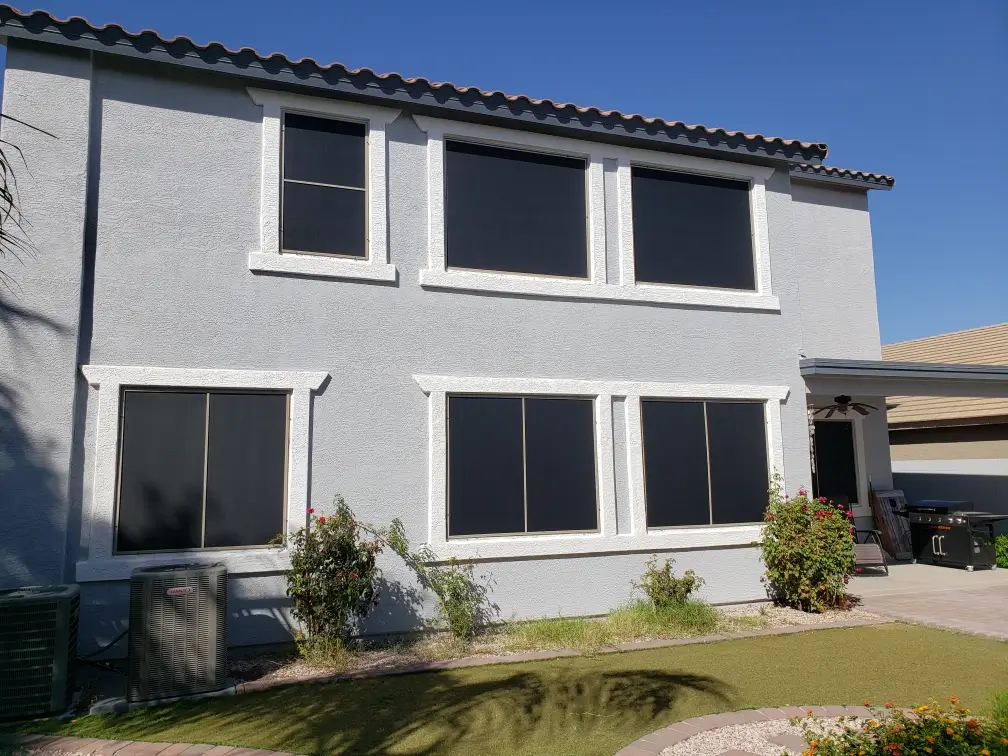 A modern two-story house with solar screens on windows, landscaped yard with greenery, and patio featuring a ceiling fan under clear blue sky.